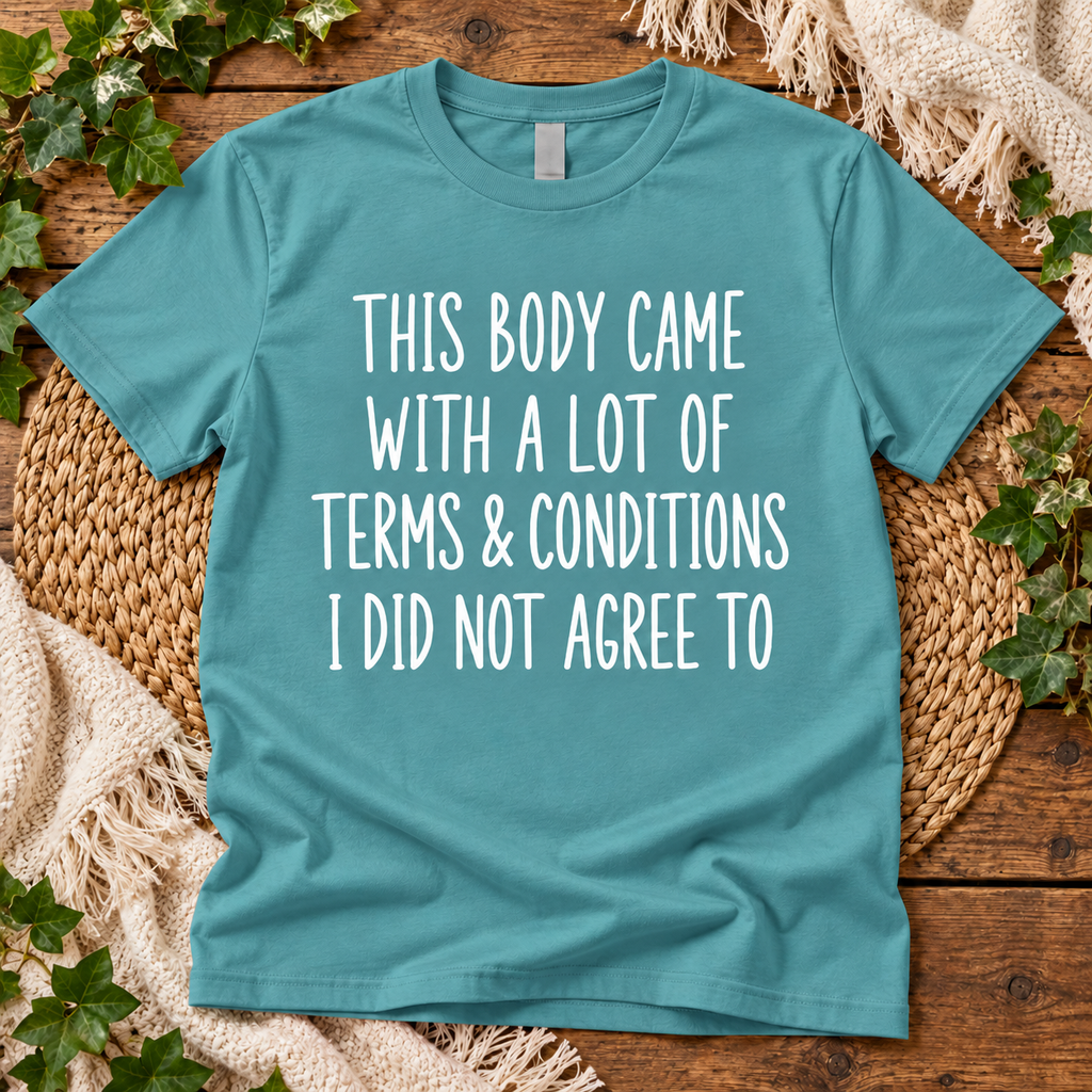 A teal short-sleeve t-shirt with white text reading 'THIS BODY CAME WITH A LOT OF TERMS & CONDITIONS I DID NOT AGREE TO', laid flat on a rustic wood surface with woven placemats, cream fabric, and green ivy around it.