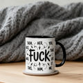 A white ceramic mug with a black handle and interior sits on a wooden surface, printed with black text and doodles. Steam rises from the mug, and a chunky gray knit blanket is draped in the background.